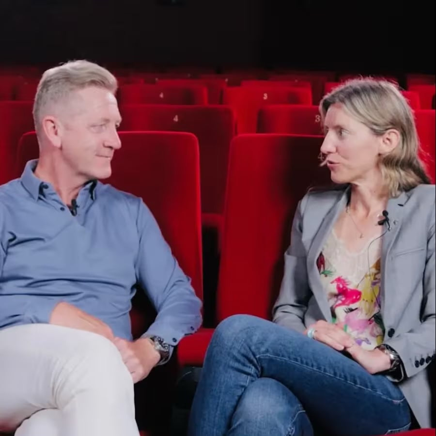 Two people sitting in a theater, engaged in conversation. Red seats in background.